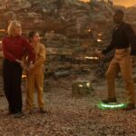 The Doctor stands on a landmine lit up green while Ruby Sunday and a little girl nervously stand in front of him in the Doctor Who episode “Boom”