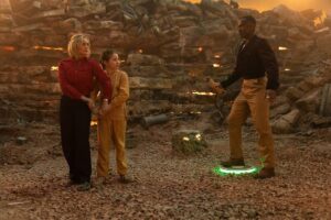 The Doctor stands on a landmine lit up green while Ruby Sunday and a little girl nervously stand in front of him in the Doctor Who episode “Boom”