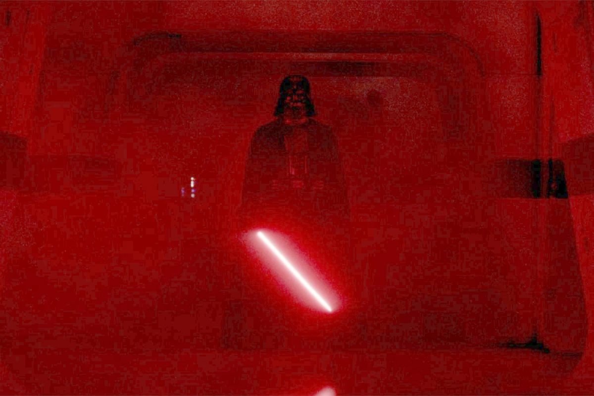 Darth Vader stands in a red washed hallway holding a sith lightsaber