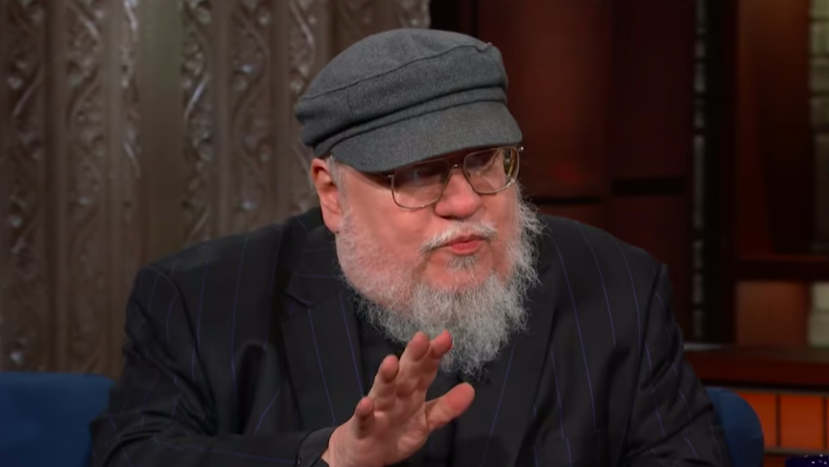George RR Martin sitting on a couch talking and wearing his trademark hat and talking to Stephen Colbert