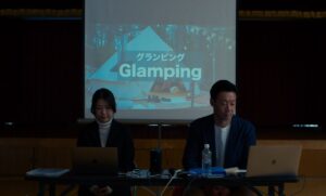 Two business people sit at a table with a slide projecting behind them that says GLAMPING in the film Evil Does Not Exist