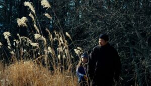 Takumi (Hitoshi Omika) walks through high, yellowing grass near the side of a forest with his young daughter Hana (Ryo Nishikawa) in Ryûsuke Hamaguchi’s Evil Does Not Exist