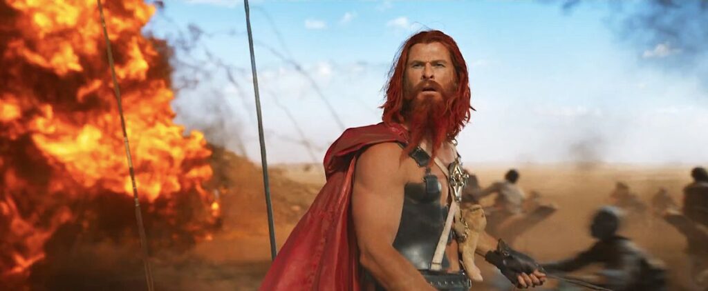 The warlord Dementus (Chris Hemsworth, in red-dyed beard and hair, with a long red cape and metal chestplate somewhat reminiscent of his MCU Thor costume, plus a teddy bear strapped to his chest) stands on a battlefield with an explosion behind him in the trailer for Furiosa