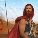 The warlord Dementus (Chris Hemsworth, in red-dyed beard and hair, with a long red cape and metal chestplate somewhat reminiscent of his MCU Thor costume, plus a teddy bear strapped to his chest) stands on a battlefield with an explosion behind him in the trailer for Furiosa