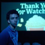 Twentysomething movie-theater employee Owen (Justice Smith) stands in a dark theater and looks at the camera, with a slide on the screen behind him that says “Thank You for Watching” atop a cartoon bucket of popcorn in Jane Schoenbrun’s I Saw the TV Glow