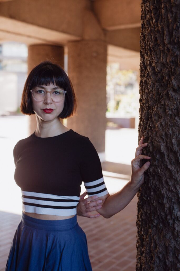 Author Kaliane Bradley stands beside a tree underneath a pavilion with an arm behind her back.