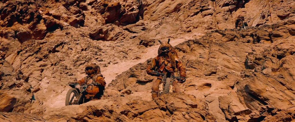 Two Rock Riders, on motorcycles, rest on a mountain face and look over the edge in Mad Max: Fury Road