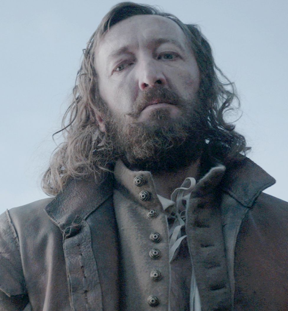 Ralph Ineson in The Witch standing in a big coat with his big beard looming over camera