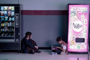 Teenagers Maddy (Brigette Lundy-Paine) and Owen (Ian Foreman) sit together on the floor in their school cafeteria, each with their back against a vending machine, in Jane Schoenbrun’s movie I Saw the TV Glow