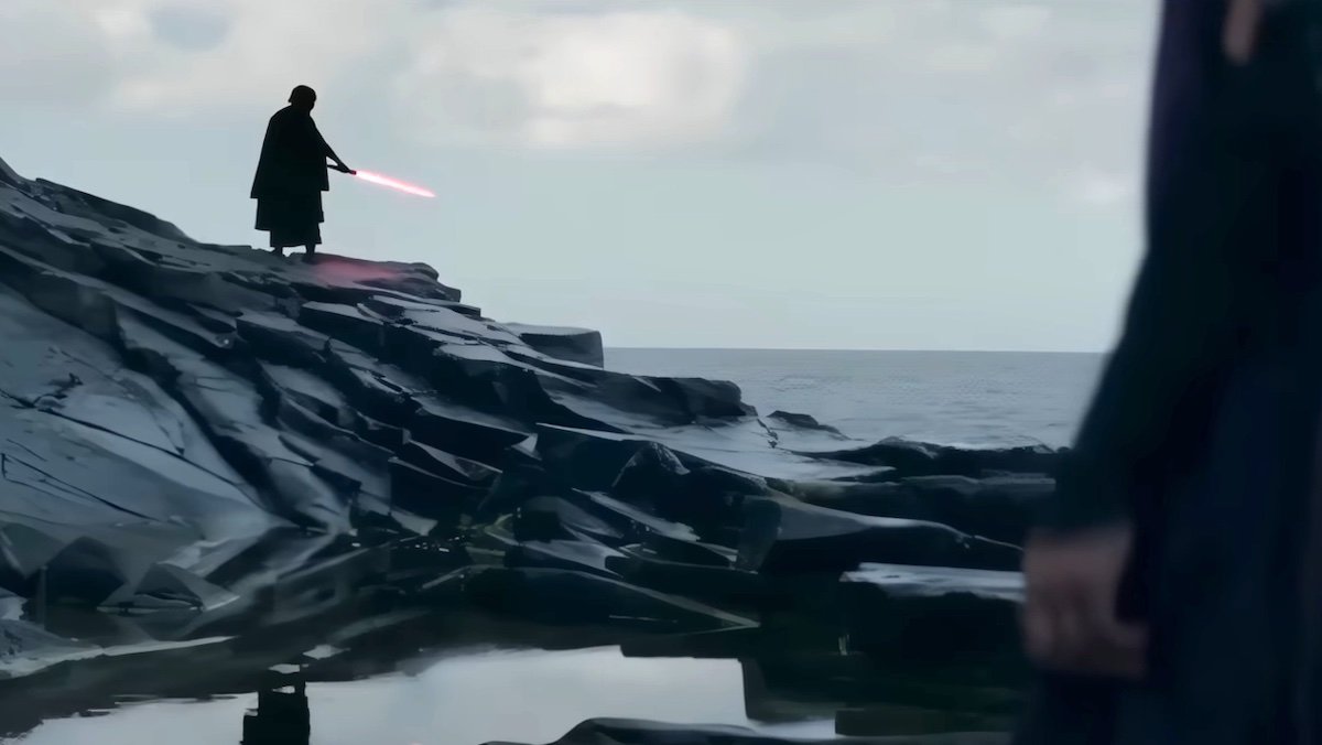 A masked figure in black holds a red lightsaber atop a rock by the ocean on The Acolyte