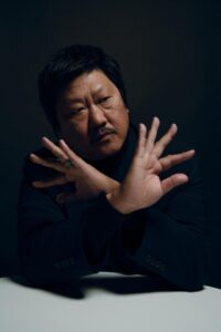 Benedict Wong rests his elbows on a table and splays his fingers wide on either side of his face