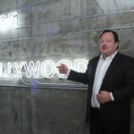 A man points to a neon "Made in Hollywood" sign on a wall.
