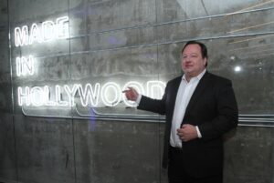 A man points to a neon "Made in Hollywood" sign on a wall.