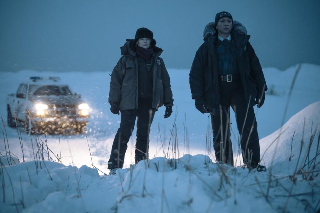 Two officers stand on a snowdrift at night, illuminated by their truck's headlights