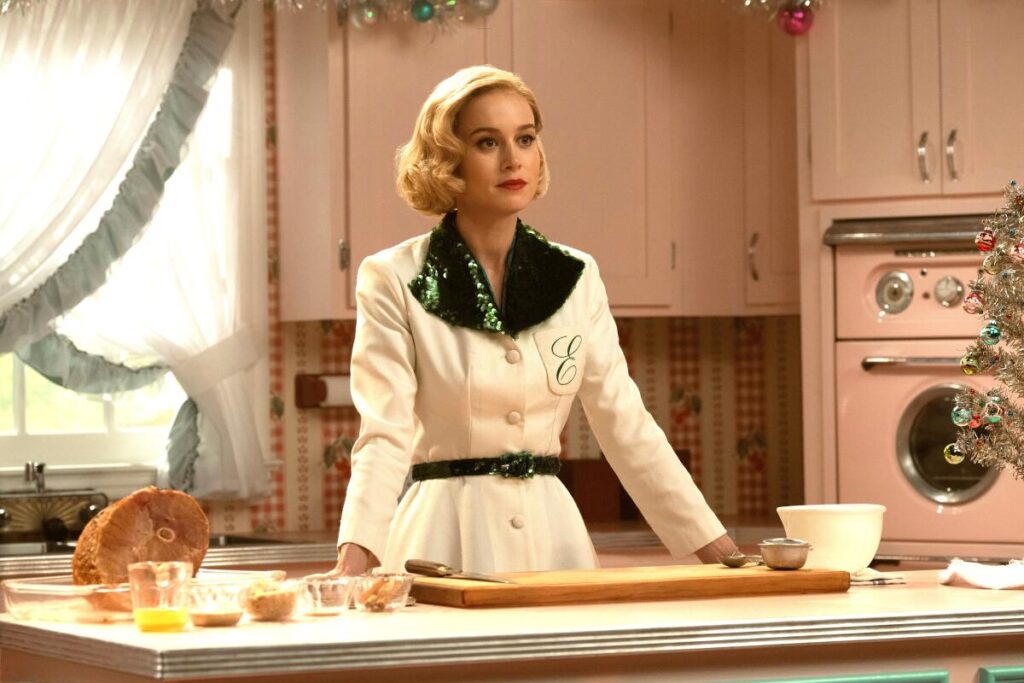 A woman in 1950s clothing stands in a pink kitchen, staring straight ahead.