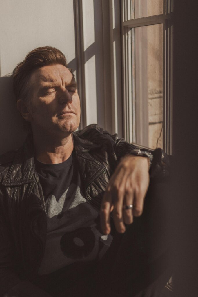 Ewan McGregor leans back against a wall with shadows from a window pane striping his face.