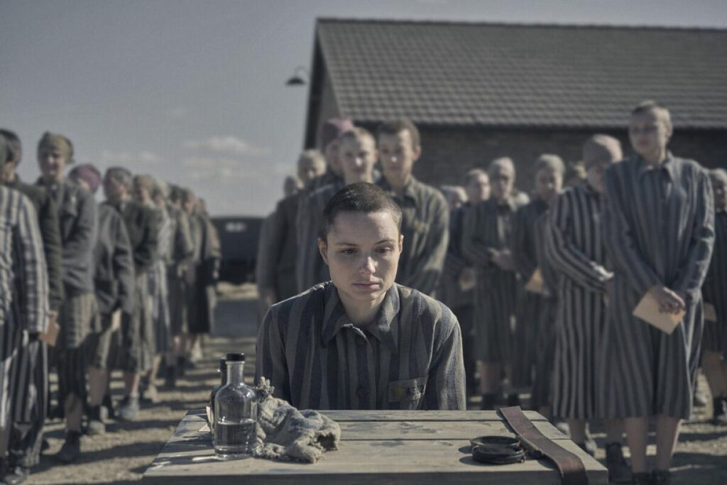 A woman camp prisoner sits at a small table with lines of other prisoners behind her in "The Tatooist of Auschwitz."