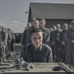 A woman camp prisoner sits at a small table with lines of other prisoners behind her in "The Tatooist of Auschwitz."