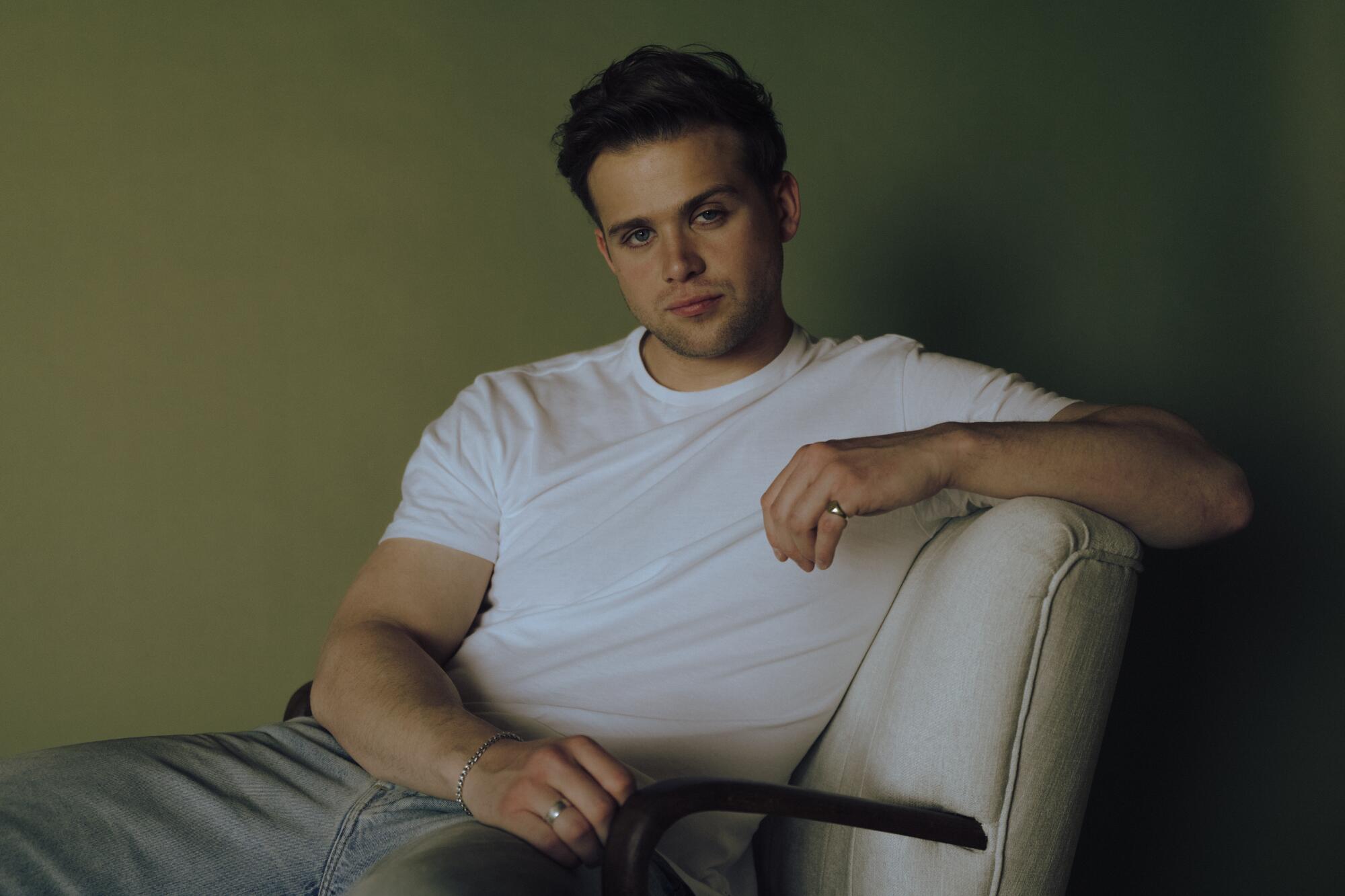 Leo Woodall in jeans and a white T-shirt, sitting in a white chair for a portrait.