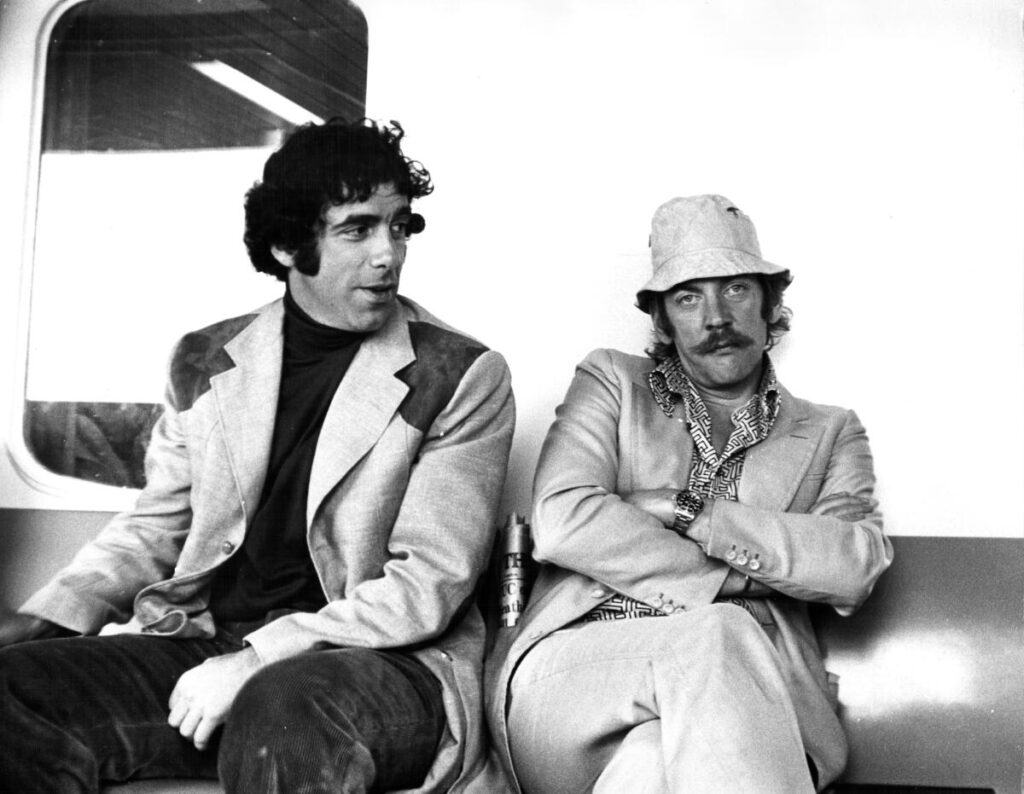 Elliott Gould looks at Donald Sutherland, with arms crossed, as both sit on a couch.