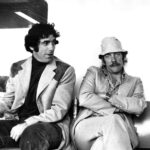 Elliott Gould looks at Donald Sutherland, with arms crossed, as both sit on a couch.