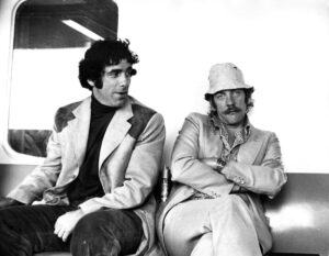 Elliott Gould looks at Donald Sutherland, with arms crossed, as both sit on a couch.