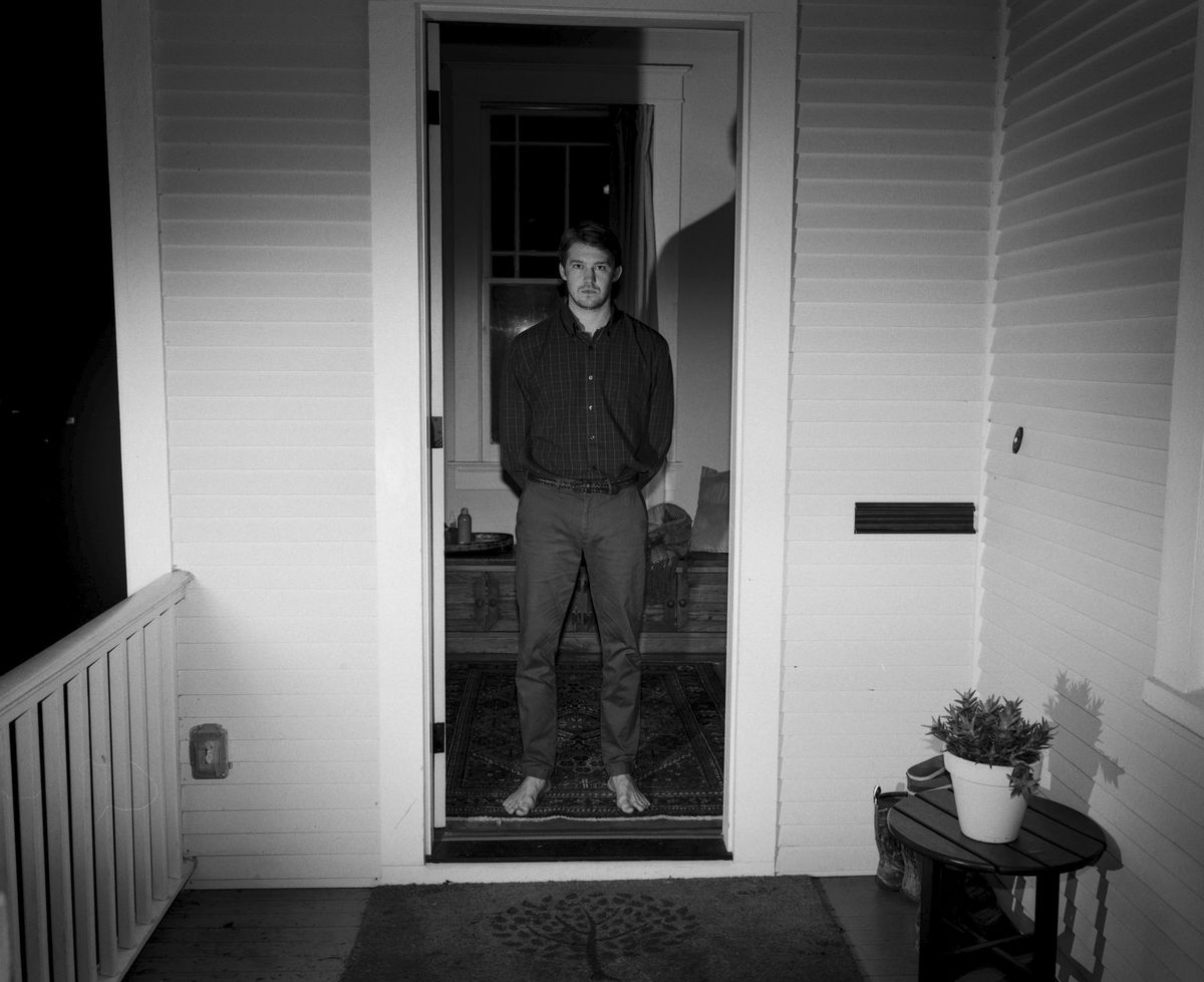 Joe Alwyn, barefoot and shot in black and white, stands in the doorway of a home in Kinds of Kindness
