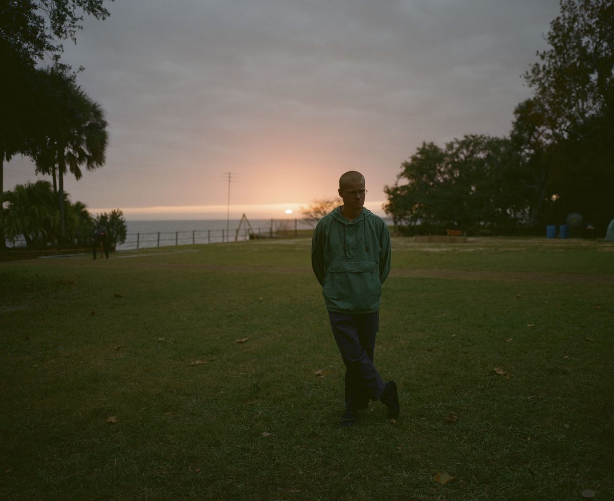 Jesse Plemons, with a buzz cut and wearing a teal windcheater, stands in a garden by the sea at sunset in Kinds of Kindness