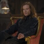 A man with long hair and dark clothing sitting on an orange velvet chair.