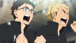 Takeda, a dark haired bespectacled man, and Ukai, a man with long dirty blonde hair, enthusiastically cheer. Both men wear matching black tracksuits.