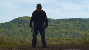 A figure with a mask on and two hooks in his hands stands facing a forest in the movie In A Violent Nature