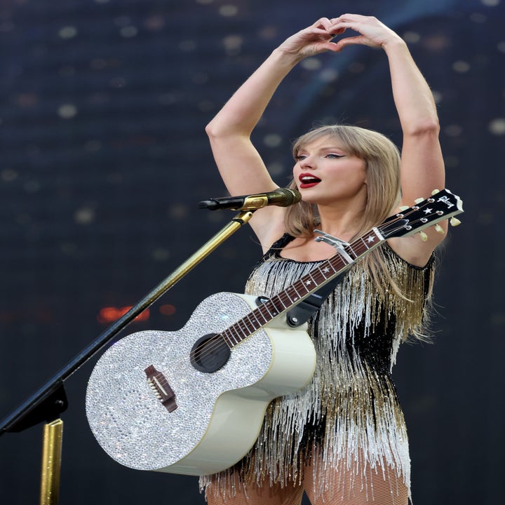 Taylor Swift on stage, wearing a fringed outfit and holding a glittery guitar, forming a heart shape with her hands above her head