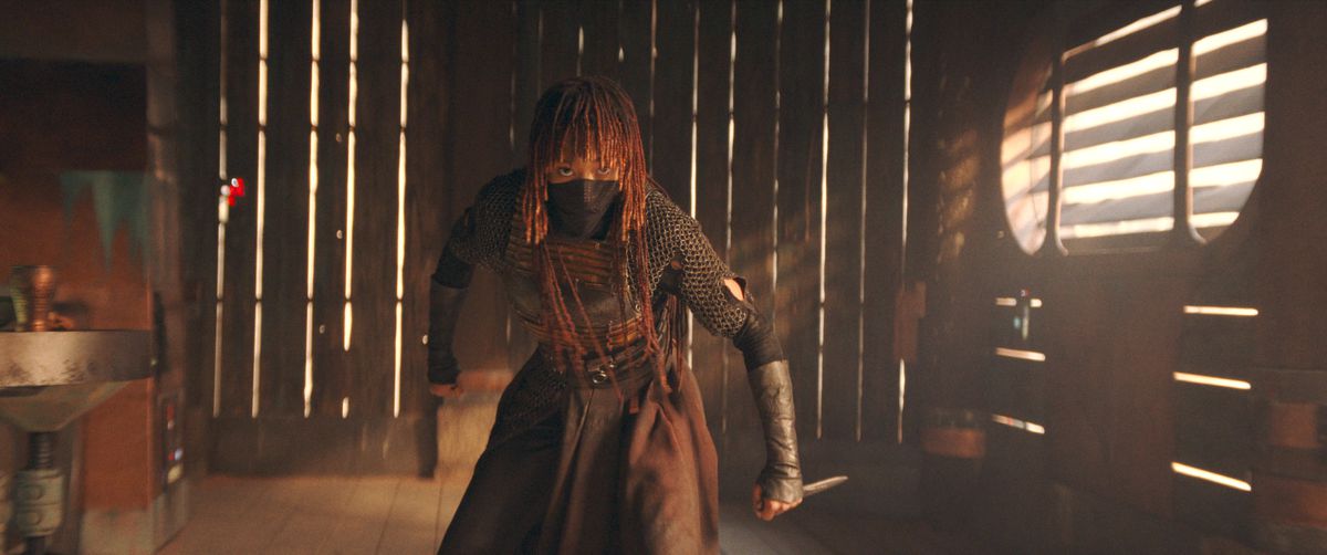 Mae (Amandla Stenberg) in The Acolyte. She’s standing in a fighting stance in a wooden-slatted building, a small dagger gripped in one hand. The bottom of her face is covered by a cloth mask.