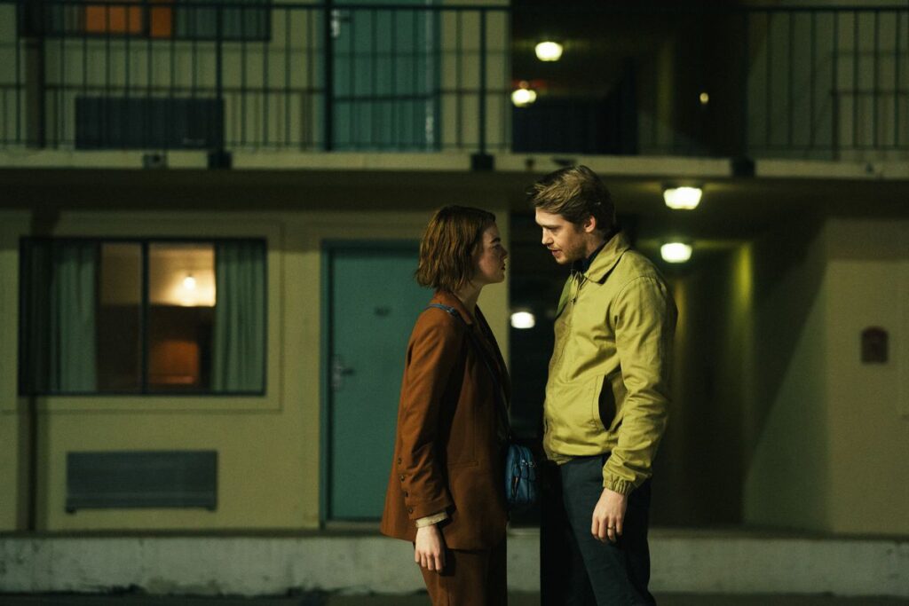 Emma Stone and Joe Alwyn stand close together in a motel car park at nighttime in Kinds of Kindness