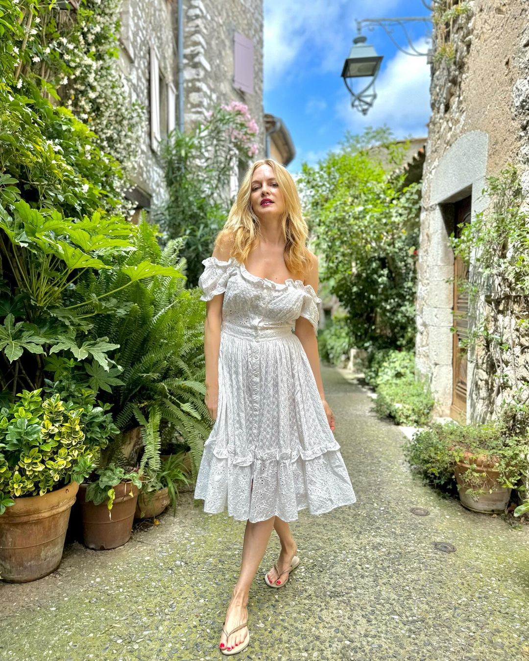 Heather Graham standing on a gorgeous path wearing a white dress and thong sandals