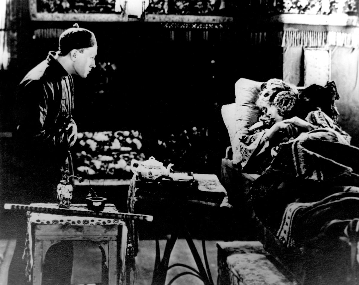 In a black-and-white film still, a seated man looks at a woman lying down