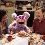 A woman and a man flank a pink and purple puppet at a dinner table.