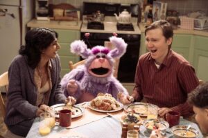 A woman and a man flank a pink and purple puppet at a dinner table.