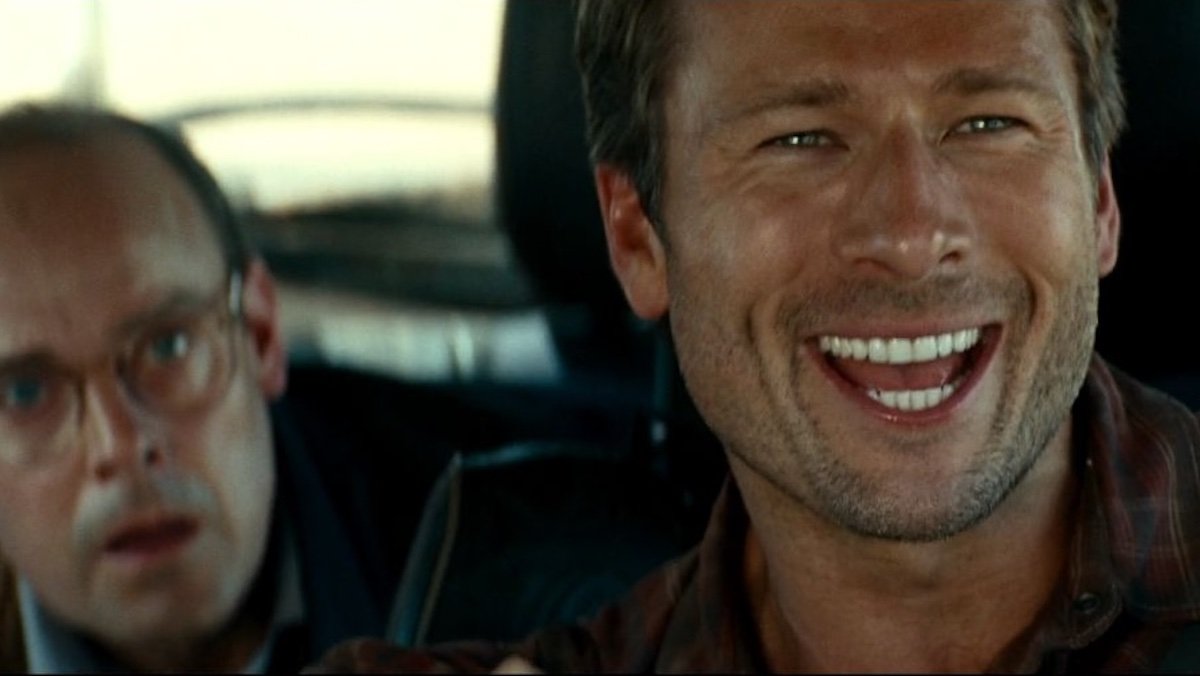 Glen Powell smiles while driving a truck as a man with glasses looks scared in the backseat in Twisters
