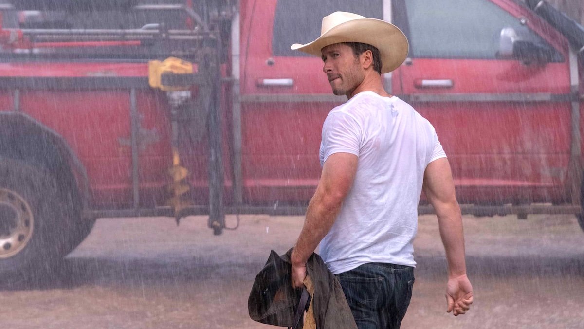 Glen POowell in a white t-shirt and cowboy hat looking back while walking in the rain towards a red truck in Twisters