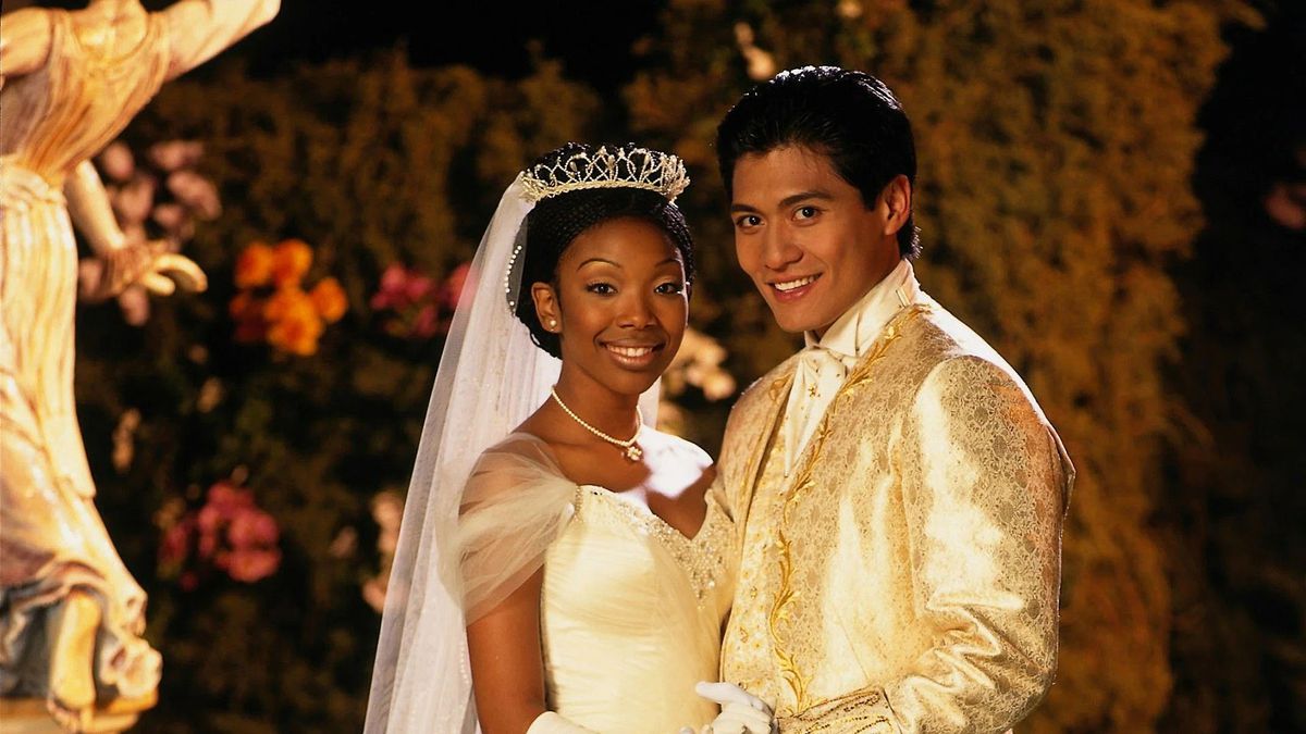 A woman in a white dress and a tiara (Brandy Norwood) and a man in a white suit (Paolo Montalban) smile in front of a background of flowers and statues.