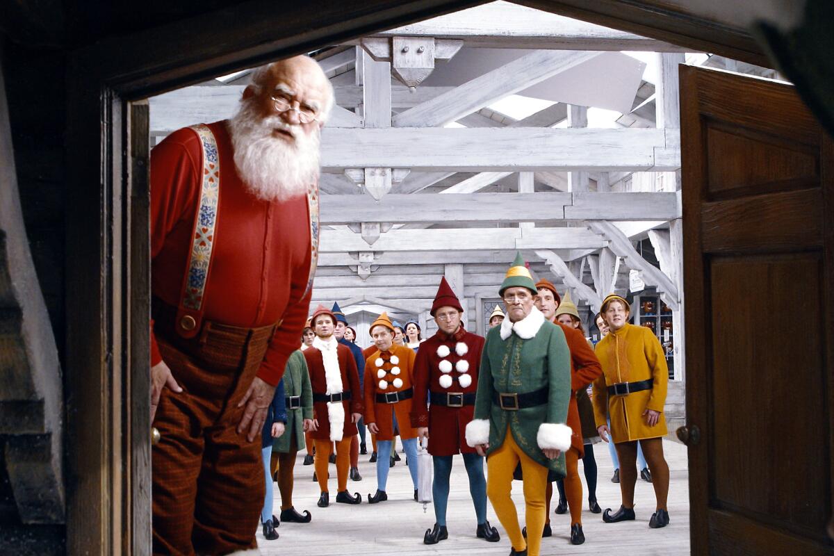 A man dressed as Santa peering through a doorway in the movie "Elf" with people dressed as elves in the background
