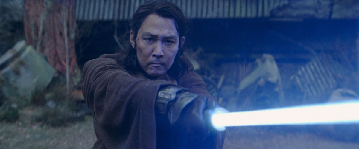 The Jedi Sol thrusts his blue lightsaber upward as he stands in the woods in a scene from The Acolyte.