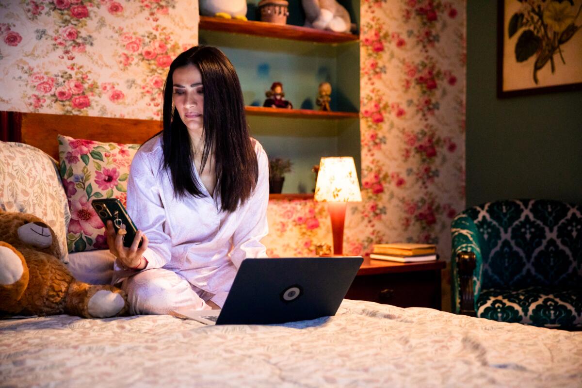 A woman in pajamas sitting on a bed with a laptop in front of her, looking at a phone in her hand.