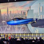 A presumably full size inflatable replica of the Fantasticar floats above the stage of Hall H at the 2024 Marvel Studios panel. The car is sleek, wheel-less, and colored in blue chrome with yellow accents.