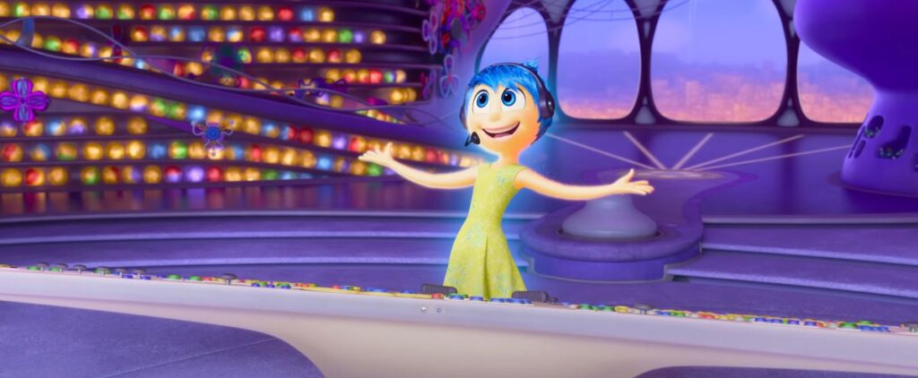 Joy from the Inside Out movies, stands at the console in emotion HQ, beaming and throwing her arms out in excitement in Inside Out 2