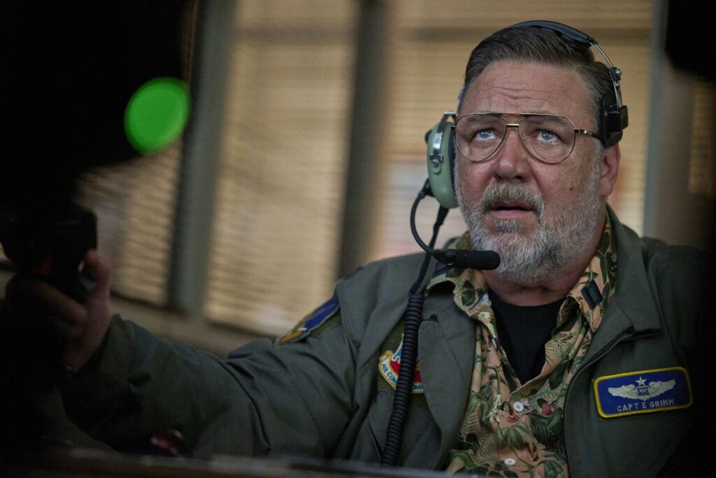 A bespectacled Russell Crowe sits in a chair and wears a headset in Land of Bad