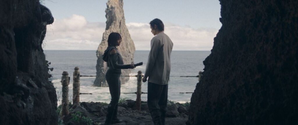 Osha and The Stranger stand in a cove overlooking the ocean, as Osha hands a lightsaber over to The Stranger in a scene from The Acolyte.
