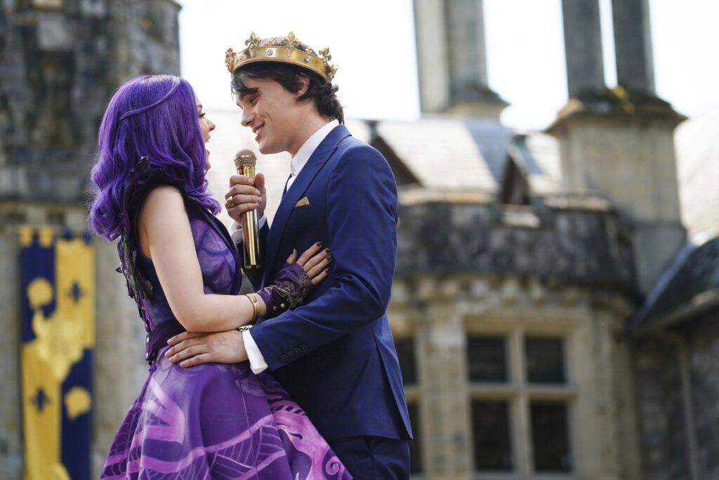 A purple haired girl in a purple ballgown looks up at a boy wearing a crown as they embrace