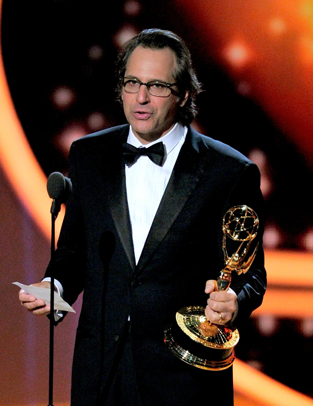 Jason Katims, in a black tuxedo, holds an Emmy statue as he delivers an acceptance speech onstage.